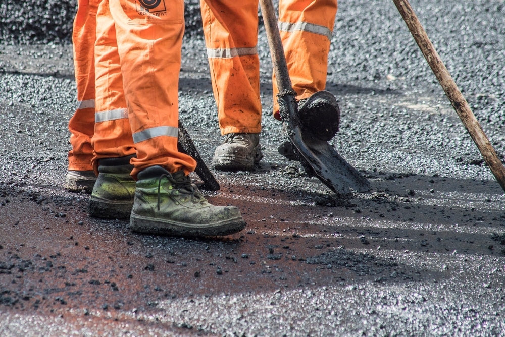 Construction de route : les bottes des ouvriers dans l'asphalte. Gros plan sur les jambes et les bottes sales d'ouvriers en pantalon orange déplaçant de l'asphalte chaud avec une pelle.