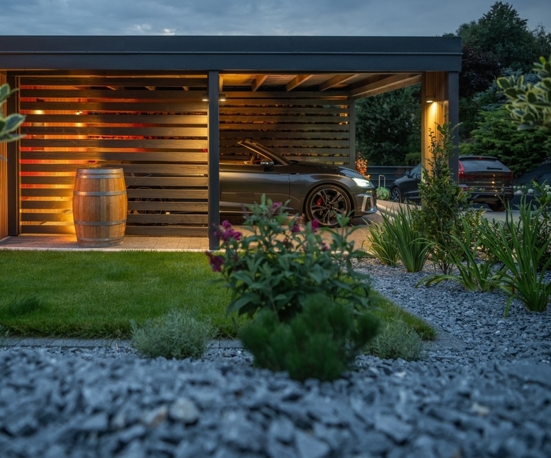 Carport bois moderne et éclairage extérieur élégant Carport moderne en bois sombre avec éclairage chaud au crépuscule. Une voiture sport et un tonneau en bois sont visibles. Jardin paysager avec gravier.
