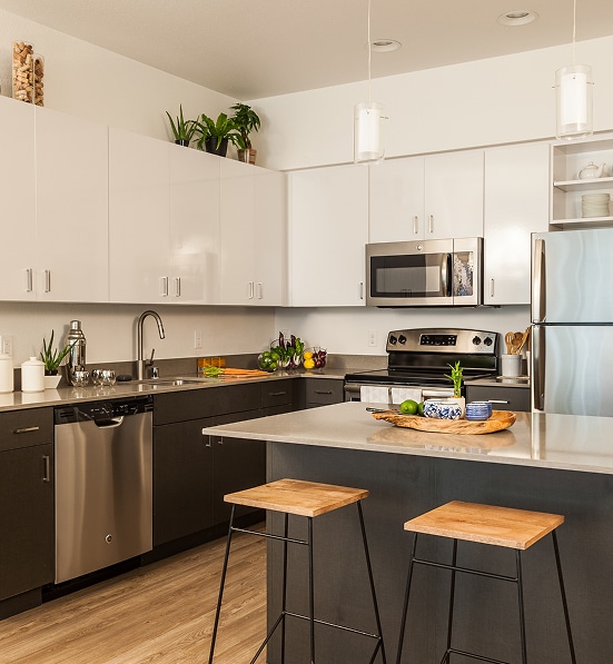 Design de cuisine moderne | Armoires blanches et îlot central. Cuisine moderne deux tons (blanc/gris foncé) en L. Îlot central avec tabourets en bois, électroménagers inox, et armoires brillantes.