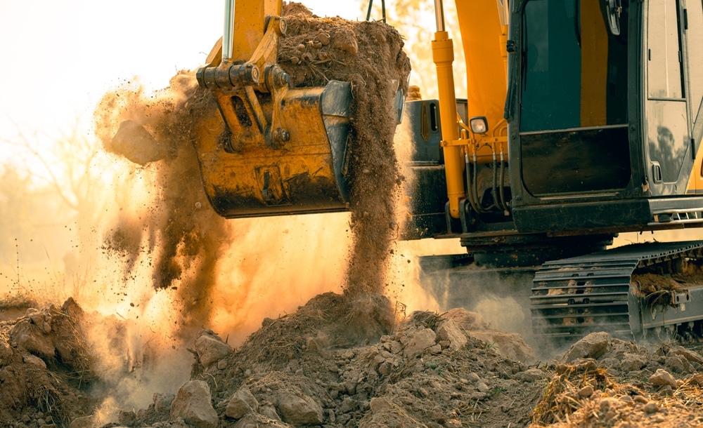Excavatrice jaune déversant terre et roches d'un godet. Poussière dorée s'élevant lors de travaux intenses sur le chantier.