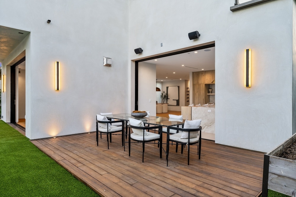 Terrasse design moderne: deck bois pour salle à manger d'extérieur. Salle à manger extérieure sur deck en bois. Murs blancs et éclairage vertical LED chaud. L'ouverture mène à une cuisine moderne.